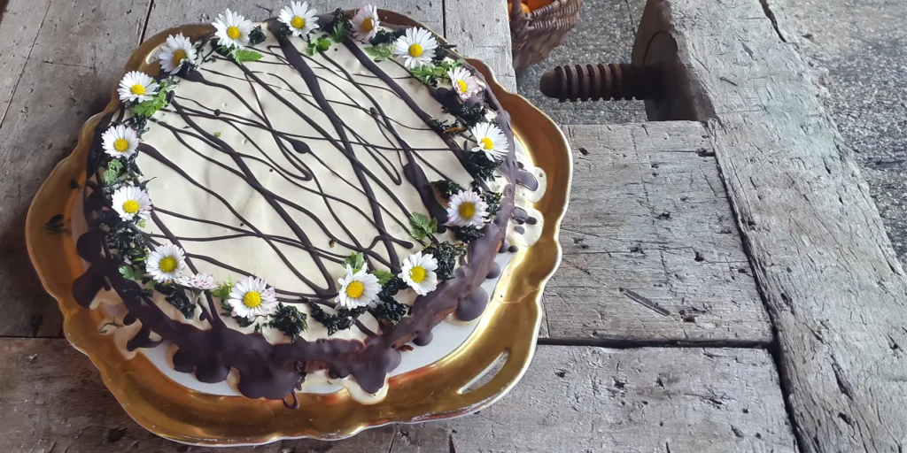 Saftiger Karotten-Orangenkuchen mit Zitronen-Cashew-Sahne-Topping geschmückt mir Gänseblümchen auf einem goldenen Servierteller auf einem rustikalen Holztisch.