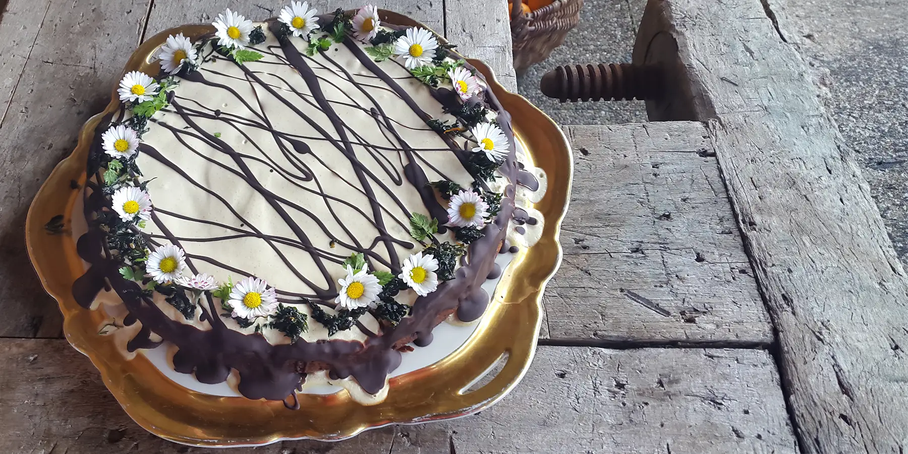 Saftiger Karotten-Orangenkuchen mit Zitronen-Cashew-Sahne-Topping geschmückt mir Gänseblümchen auf einem goldenen Servierteller auf einem rustikalen Holztisch.