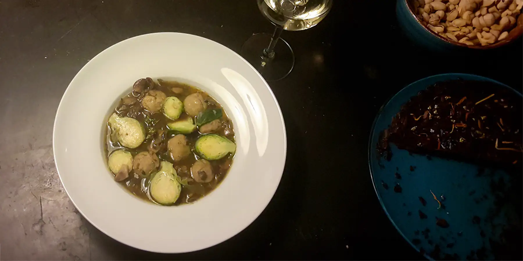 Ein weißer Teller mit Wald- & Wiesenpilzsuppe mit Grünkernklößchen von oben fotografiert neben einem Glas Weißwein.