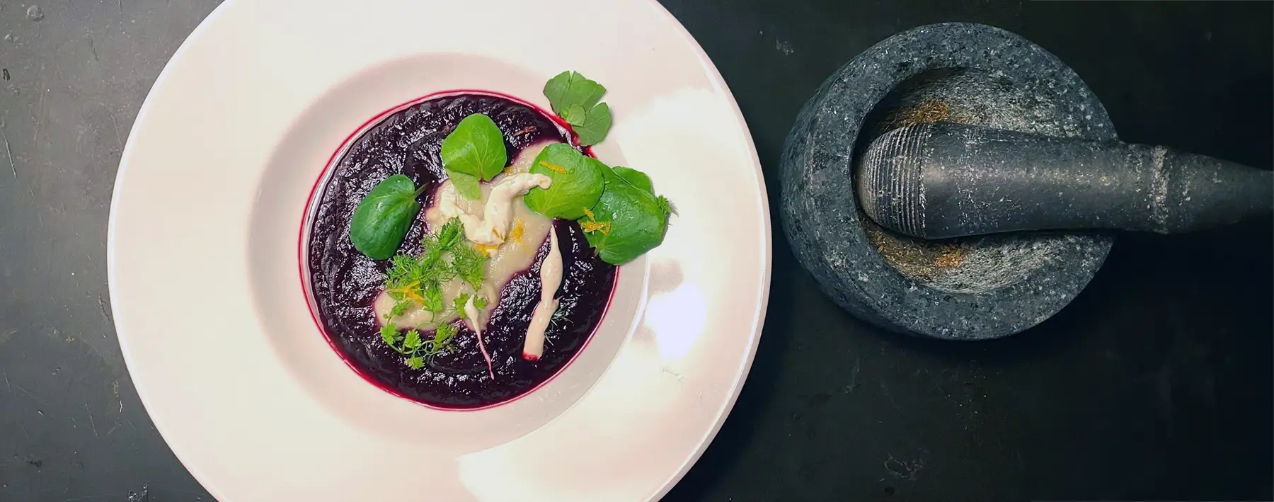 Ein weißer Teller gefüllt mit Rote-Beete-Holunder-Suppe mit Fenchel-Schaum und Cashnew-Sahne neben einem Mörser mit Stößel aus Granit.