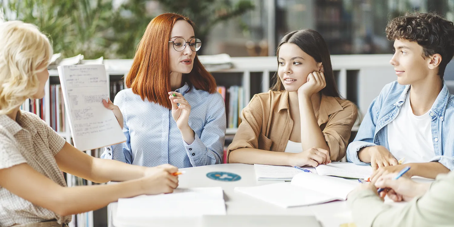 Eine Gruppe an Teenagern sitzen im Austausch mit ihrer Vertrauensperson an einem Tisch als Symbol für den MHFA Ersthelfer-Kurs Teen.